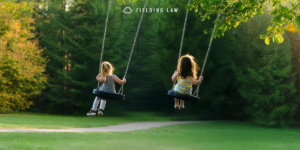 two young female friends playing in a backyard on swings with trees in the background.