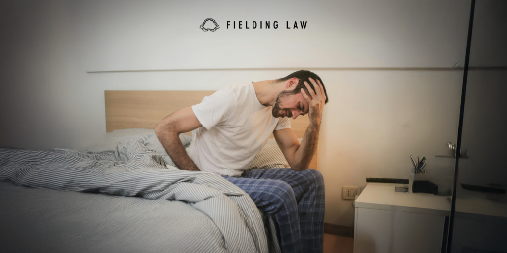 Man sitting at the edge of a bed touching his back and head from an injury