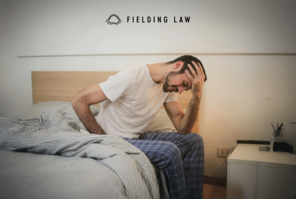 Man sitting at the edge of a bed touching his back and head from an injury