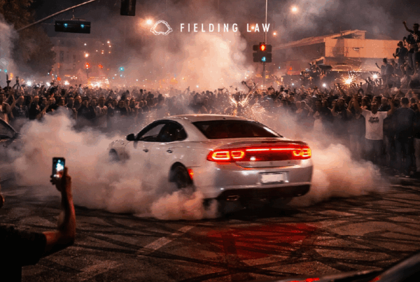 A white care in the middle of the street intersection with tire smoke and a large crowd of people around. Street takeover at nightfall